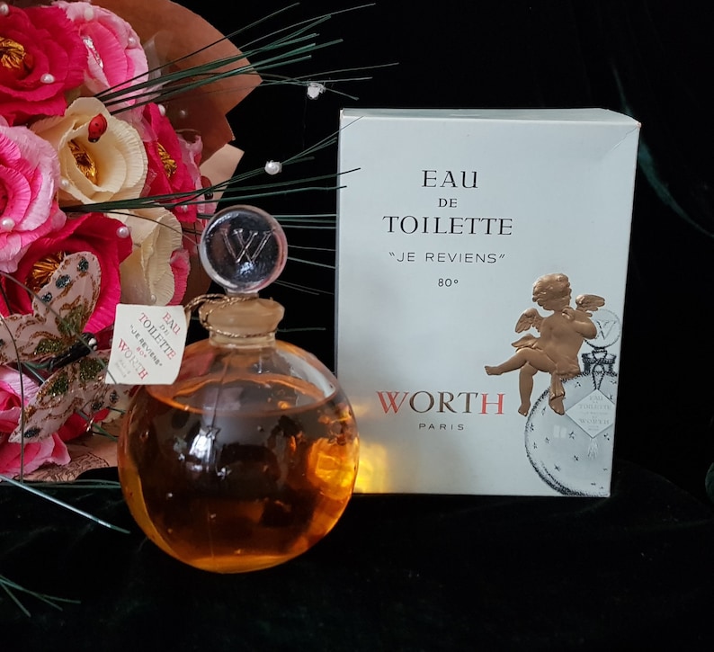 May include: A vintage glass bottle of perfume with a gold cap and a gold label that reads "Eau de Toilette "Je Reviens" 80&deg; Worth Paris". The bottle is round and clear, and the liquid inside is a light amber color. The bottle is sitting on a black surface.