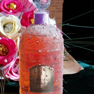 May include: A clear glass bottle with a purple ribbon and a decorative stopper. The bottle is filled with a pink liquid and has a gold label that says "Instant Real". The bottle is surrounded by a bouquet of pink and white flowers.