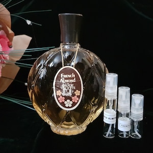 May include: A clear glass bottle of French Almond Eau de Toilette with a textured, diamond-shaped design. The bottle has a brown label with the text "French Almond Eau de Toilette". Three small clear glass spray bottles are in front of the larger bottle.