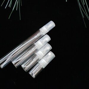 May include: Four clear glass travel-sized spray bottles with white caps. The bottles are arranged in a pyramid shape on a black background.