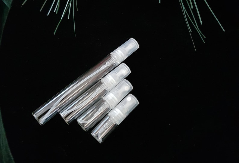 May include: Four clear glass travel-sized spray bottles with white caps. The bottles are arranged in a pyramid shape on a black background.