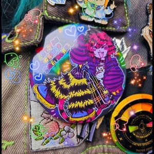 Op de afbeelding: Een ronde knoop met een holografische afwerking met een anime-achtig personage met roze haar en een geel en zwarte outfit. De knoop heeft de tekst "LET'S BE CRINGE TOGETHER!!" en is omringd door andere decoratieve spelden.