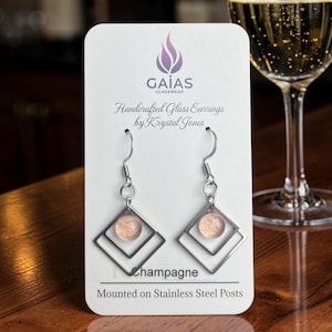 Peut inclure: Une paire de boucles d'oreilles sur un présentoir blanc marqué GAIAS GLASSWEAR Handcrafted Glass Earrings by Krystal Jones. Les boucles d'oreilles présentent des cadres carrés en métal argenté avec un insert en verre texturé couleur champagne.