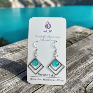 Peut inclure: Une paire de boucles d'oreilles pendantes avec une petite perle de verre ronde turquoise centrée dans deux cadres carrés argentés imbriqués. Les boucles sont fixées sur une carte blanche marquée GAIAS GLASSWEAR, Handcrafted Glass Earrings by Krystal Jones, Moraine Lake, et Mounted on Stainless Steel Posts.