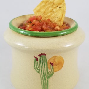 May include: A ceramic salsa dish with a green rim, featuring a painted cactus and a sun design. The dish is filled with salsa and topped with a tortilla chip.