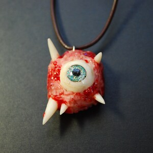 May include: A necklace with a pendant shaped like a bloody eye with white spikes. The eye has a blue and green iris with a black pupil.