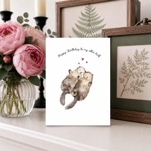May include: A white greeting card featuring a watercolor illustration of two brown otters floating together. Above the otters are three small pink hearts and the black text that reads, Happy Birthday to my otter half, centered on the card.