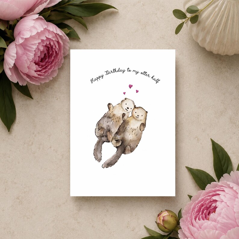 May include: A white greeting card featuring a watercolor illustration of two brown otters holding hands. Above the otters are three small pink hearts and the black text "Happy Birthday to my otter half" written in a simple, rounded font.