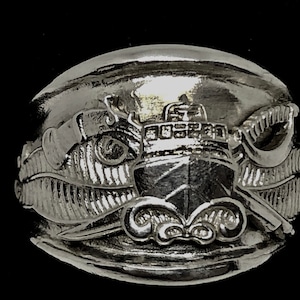 May include: A silver-toned ring featuring a detailed ship design. The ring showcases a ship with intricate detailing, waves, and a sword. The ring is set against a black background, highlighting the metallic sheen and the nautical theme.
