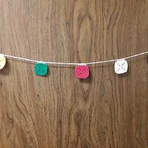 Puede incluir: Una cadena de cuadrados de ganchillo en amarillo, verde, rojo, blanco y azul colgando de un fondo de madera.