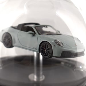 Könnte beinhalten: Hellgraues Cabrio-Sportwagenmodell mit schwarzen Rädern und roten Bremssätteln. Das Modell wird in einer transparenten Glaskuppel präsentiert. Das Auto hat das Wort "GTS" auf der Seite.