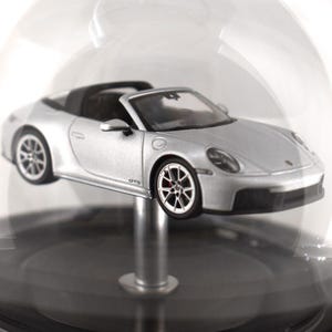Könnte beinhalten: Silbernes Cabrio-Sportwagenmodell mit schwarzem Innenraum und Rädern. Das Modell wird in einer transparenten Glaskuppel auf einem schwarzen Sockel ausgestellt. Das Auto hat das Wort "GTS" an der Seite.