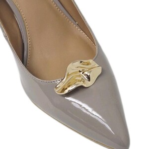 May include: Close-up of a gray patent leather pump with a pointed toe. A gold-colored decorative accent is affixed to the vamp. The shoe's interior is a light tan color.