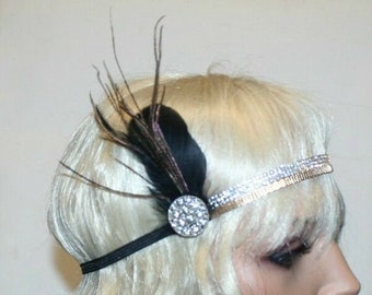 May include: A black and gold headband with a rhinestone embellishment and a black feather accent. The headband is worn on a mannequin with blonde hair.