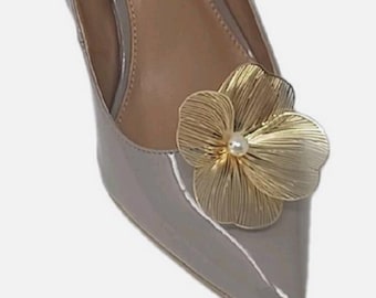 May include: A gray patent leather pump with a pointed toe. A gold-colored metal flower with a pearl center adorns the vamp. The shoe has a high heel and a neutral-colored insole.