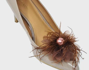 May include: A beige patent leather pointed-toe pump with a stiletto heel. The shoe features a decorative embellishment on the toe consisting of brown feathers, sheer brown fabric petals, and a single glossy pink pearl-like bead in the center.
