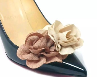 May include: Close-up of a black patent leather high-heeled shoe adorned with two fabric flower embellishments. One flower is a light beige color, and the other is a muted rose color. The shoe has a pointed toe and a glossy finish.