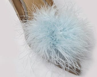 May include: Close-up of a silver-colored pointed-toe shoe adorned with a fluffy, light blue feather embellishment. The shoe's interior is a light tan color, and the background is white.