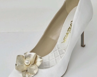 May include: A white satin pointed-toe pump featuring a slim stiletto heel and a quilted fabric detail on the inner side. The shoe is adorned with a metallic gold flower embellishment at the toe, centered with a single white pearl accent.