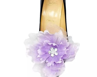 May include: Black patent leather high heel shoe with a large, layered lavender and white fabric flower accent. The flower has a sparkling, rhinestone center. The shoe's sole is gold with the text "Paris".