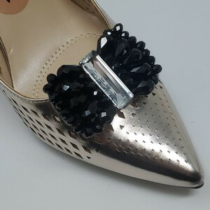 May include: Gold-colored shoe with a black beaded bow and a clear crystal accent.
