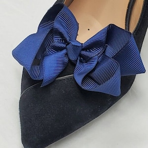 May include: Close-up of a navy blue bow attached to a black suede shoe. The bow is made of a ribbed fabric and is centered on the shoe's toe. The shoe has a pointed toe and a low heel, suggesting a formal or dressy style.