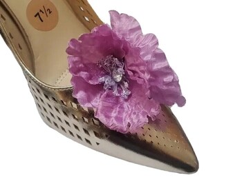 May include: A metallic gold pointed-toe pump with a laser-cut diamond pattern and a large, ruffled purple fabric flower attached to the toe. The interior insole features the text 7 1/2 printed on a circular tan sticker.