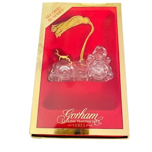 May include: A clear glass Christmas ornament depicting a vintage car with a gold reindeer on the hood. The ornament is in its original box, which is red with gold trim and features the Gorham logo and the text "A Holiday Tradition Since 1821".