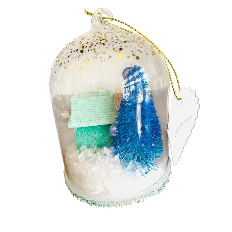 May include: A clear glass Christmas ornament with a miniature winter scene inside. The scene includes a teal house, a blue bottle brush tree, and faux snow. The ornament is decorated with gold glitter and has a gold string for hanging.