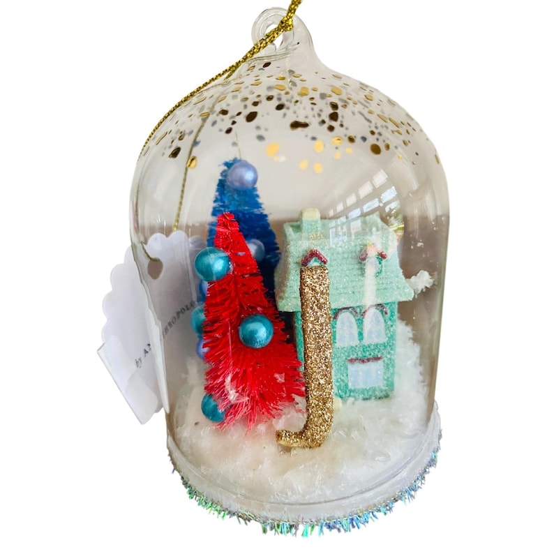May include: A clear glass Christmas ornament with a miniature winter scene inside. The scene includes a red and blue tree, a green house, and faux snow. The ornament is decorated with gold glitter and a gold hanging string.