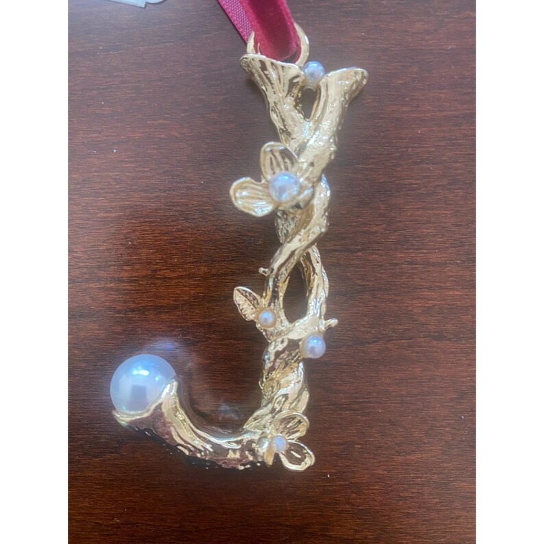 Puede incluir: Un adorno dorado con la letra "J" y un acento de perla. La letra est&aacute; dise&ntilde;ada con un motivo floral y de vid, adornada con peque&ntilde;as perlas y una cinta burdeos para colgar.