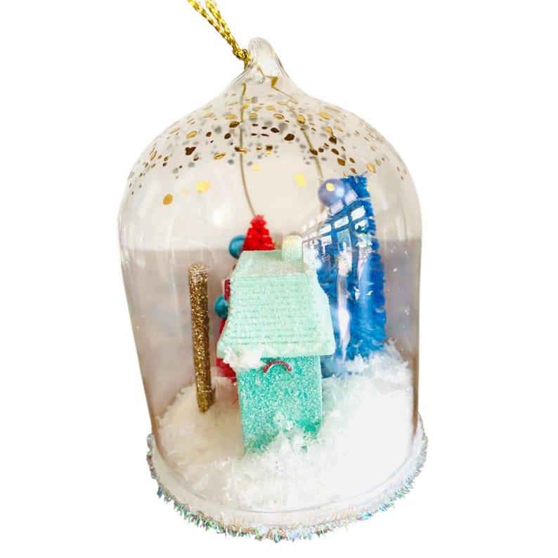 May include: A clear glass Christmas ornament shaped like a dome, with a miniature winter scene inside. The scene includes a teal house, a blue tree, and a gold glitter pole, all set in a snowy landscape. The ornament is decorated with gold glitter.