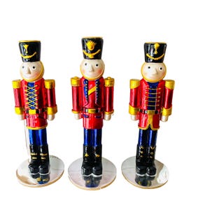 May include: Three decorative toy soldier figurines. Each soldier is dressed in a red jacket with gold accents, blue pants, and a black hat. The figurines stand on silver circular bases, and have smiling faces.