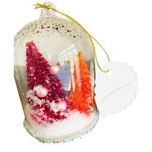 May include: A clear glass Christmas ornament with a gold string. Inside, there are miniature pink and orange bottle brush trees, pink ornaments, and a small blue house. The base is white with iridescent trim. A white tag reads "by ANTHROPOLOGIE".