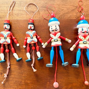 May include: Four wooden toy soldier ornaments with red, white, blue, and black paint. Each has a string for hanging, with articulated arms and legs. The ornaments are on a brown wooden surface.