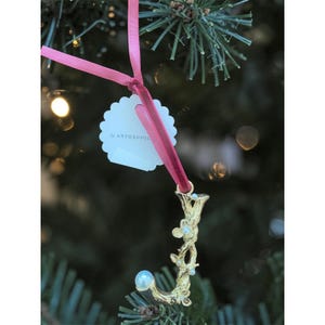 Puede incluir: Adorno navide&ntilde;o dorado en forma de la letra "J" con detalles florales y perlas, colgado de una cinta rosa. El adorno tiene una etiqueta que dice "by ANTHROPOL". El fondo es un &aacute;rbol de Navidad borroso.