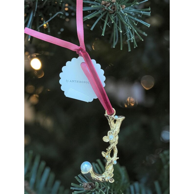 Puede incluir: Adorno navide&ntilde;o dorado en forma de la letra "J" con detalles florales y perlas, colgado de una cinta rosa. El adorno tiene una etiqueta que dice "by ANTHROPOL". El fondo es un &aacute;rbol de Navidad borroso.