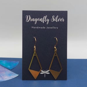 May include: A pair of gold-toned geometric drop earrings with a diamond-shaped cutout design, suspended from gold-toned hooks. The earrings are displayed on a dark blue card featuring the text Dragonfly Silver Handmade Jewellery.