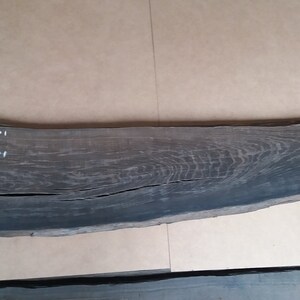 May include: A dark grey wood slab with a rough, natural edge. The slab is 58 inches long, 13 inches wide, and 1.6 inches thick.