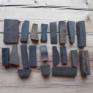 May include: An assortment of small, dark gray wooden blocks in various shapes and sizes, arranged on a light brown wooden surface. The blocks include rectangular, curved, and angled pieces, showcasing natural wood grain.