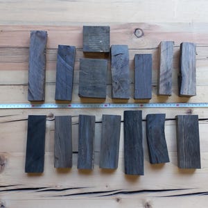 May include: An overhead shot displays an assortment of small, rectangular wooden blocks in varying shades of gray and black. A measuring tape is laid across the wooden blocks. The blocks are arranged on a light brown wooden surface.