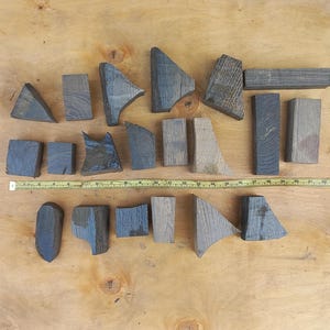 May include: A collection of twenty-one dark, weathered wooden blocks of various geometric shapes and sizes arranged in rows, accompanied by a yellow measuring tape that spans approximately 60 centimetres across the centre of the display.