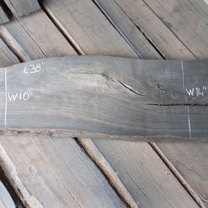 Op de afbeelding: Een donkergrijze houten plaat met een natuurlijke rand, 38 inch lang en 10 inch breed. Het hout heeft een knoop en een scheur in het midden.