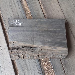 May include: A rectangular piece of dark gray wood with the dimensions 13" x 8". The wood has a rough, textured surface with visible grain and a weathered appearance. The edges show signs of natural wear and tear.