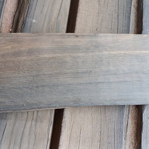 May include: A long, narrow piece of wood with a gray stain. The wood has a rough, natural texture and a slightly uneven edge.