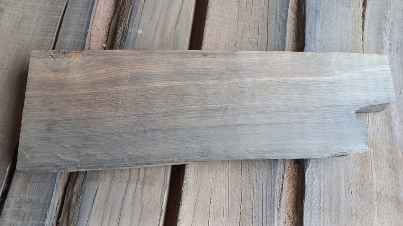 May include: A long, narrow piece of wood with a gray stain. The wood has a rough, natural texture and a slightly uneven edge.