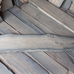 May include: A large piece of rough-hewn wood with a natural edge. The wood is dark brown and grey in colour. The wood is 64 inches long, 12 inches wide, and 1.8 inches thick. The measurements are written in white chalk on the wood.