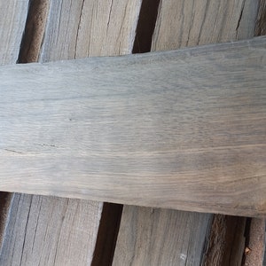 May include: A long, rectangular piece of wood with a gray-brown color and a natural wood grain pattern. The wood has a rough, unfinished texture.