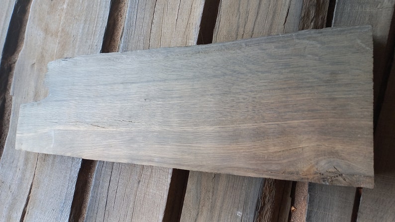 May include: A long, rectangular piece of wood with a gray-brown color and a natural wood grain pattern. The wood has a rough, unfinished texture.