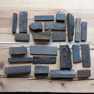 May include: An assortment of weathered, dark gray wooden blocks of varying shapes and sizes, arranged on a light brown wooden surface. A measuring tape is laid across the blocks. The blocks appear to be reclaimed or repurposed wood.